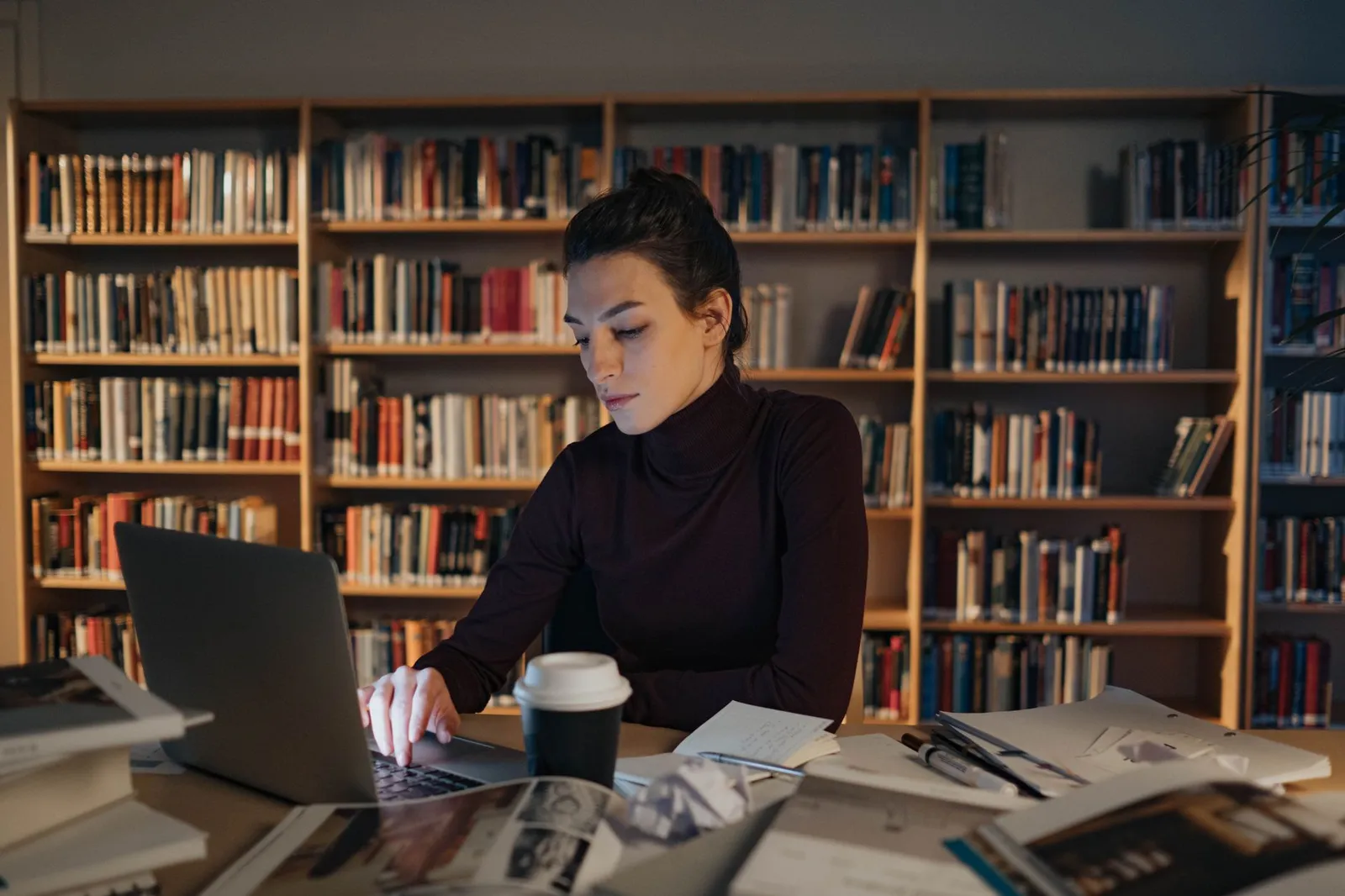 Woman typing on a laptop at a library table, researching online AI degrees