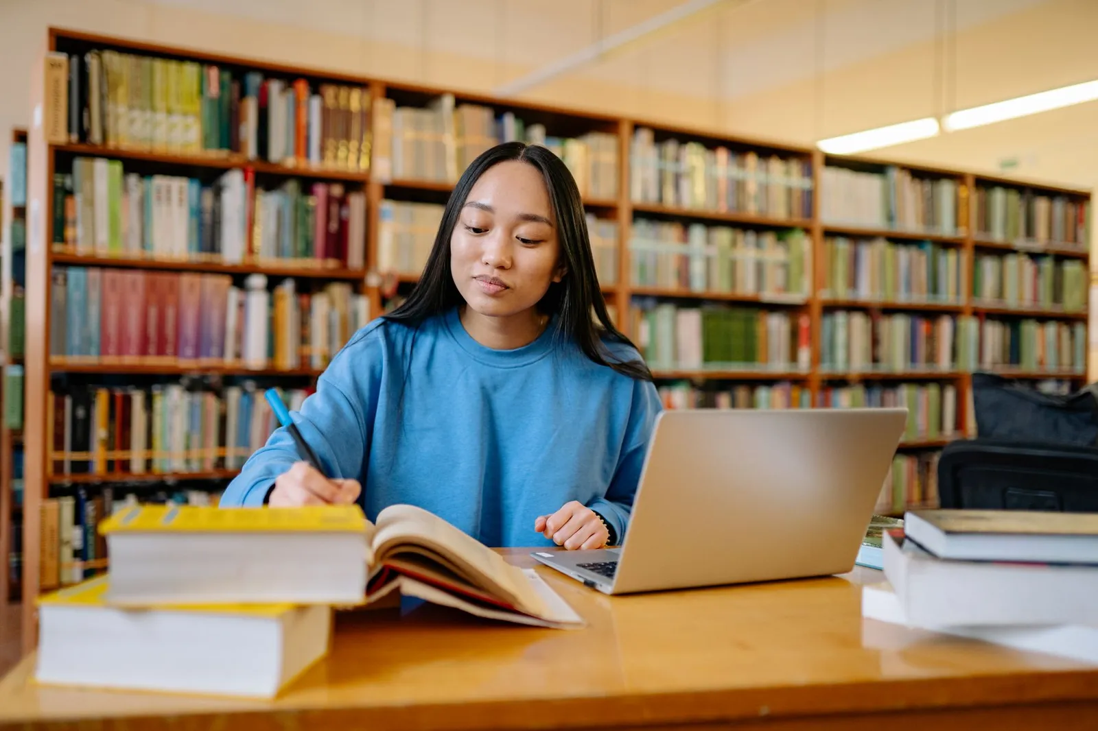 Woman taking notes from a textbook beside a laptop in a library, studying artificial intelligence certificate programs