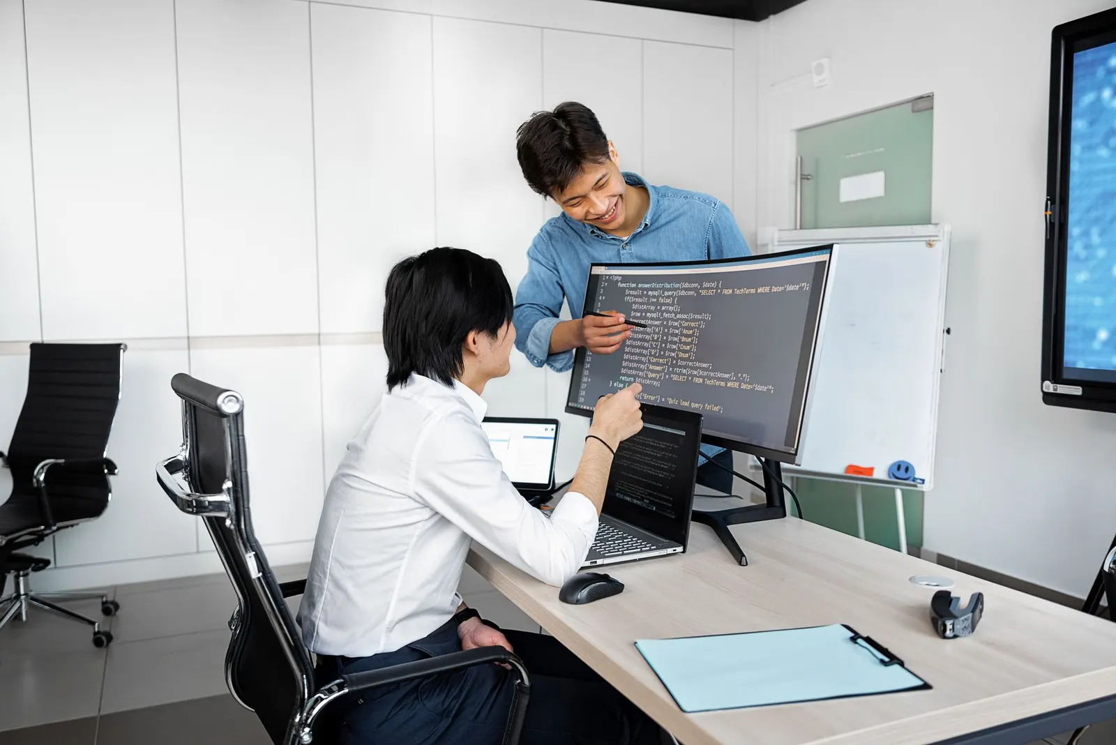 AI engineer pointing at code on a curved monitor as a colleague reviews it at the desk