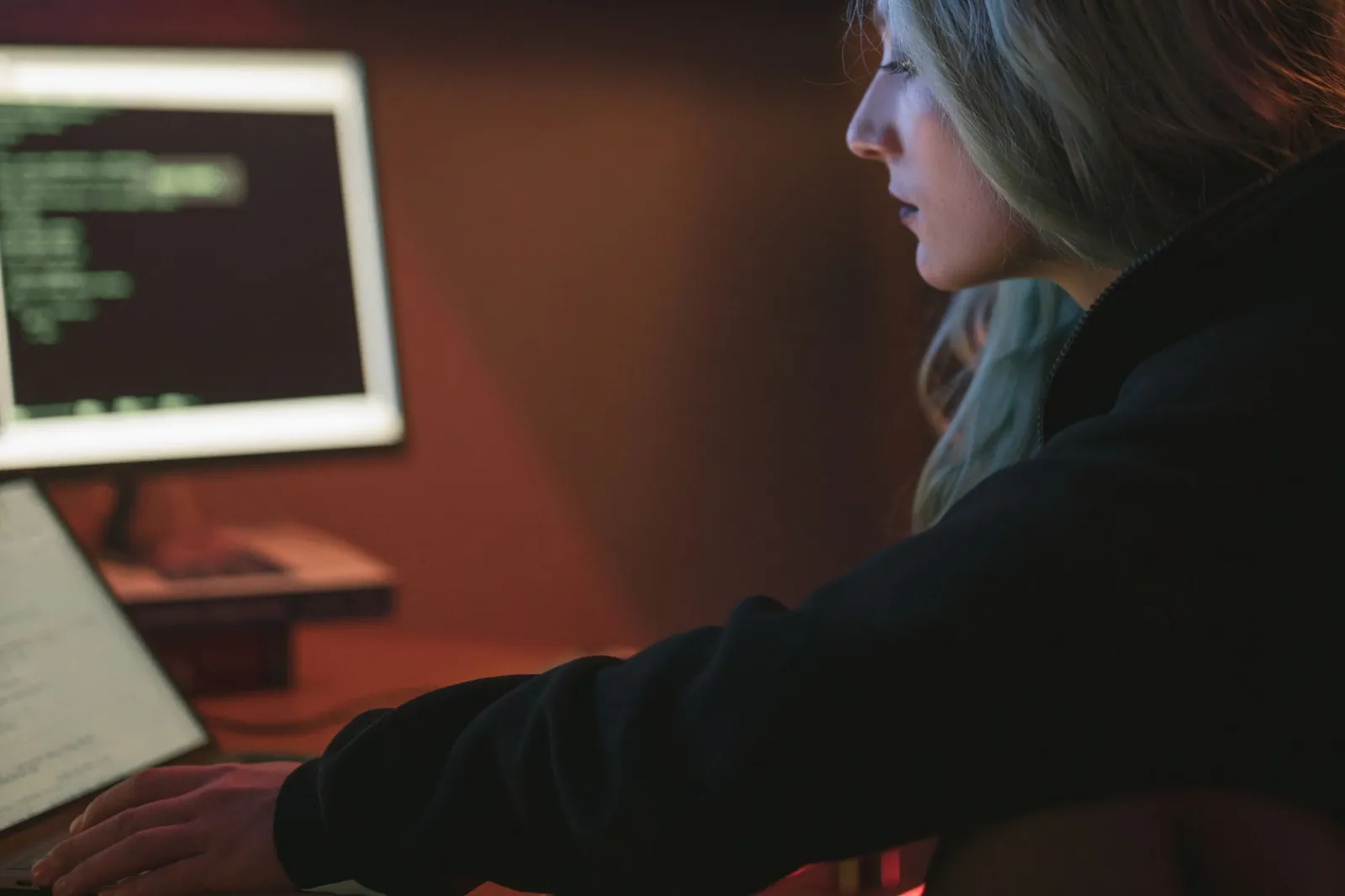 Woman working late at a laptop beside a monitor of green text, studying for AWS machine learning specialty