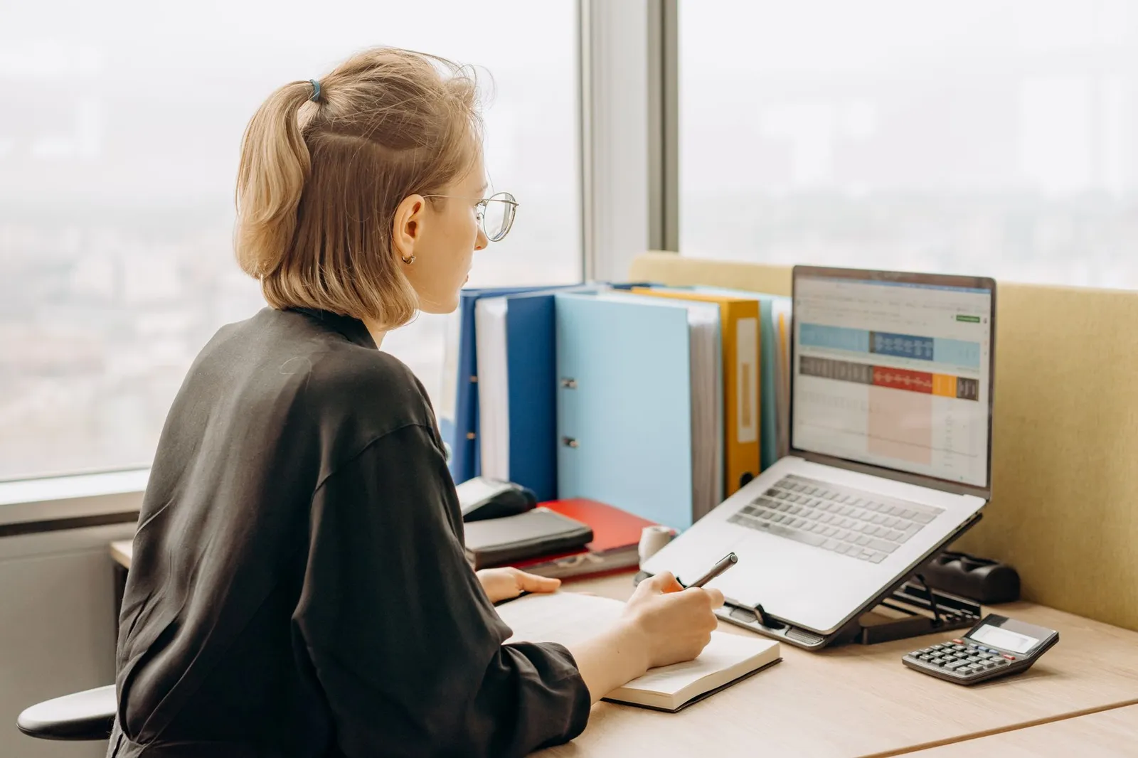 Data analyst taking notes beside a laptop spreadsheet at a desk by the window