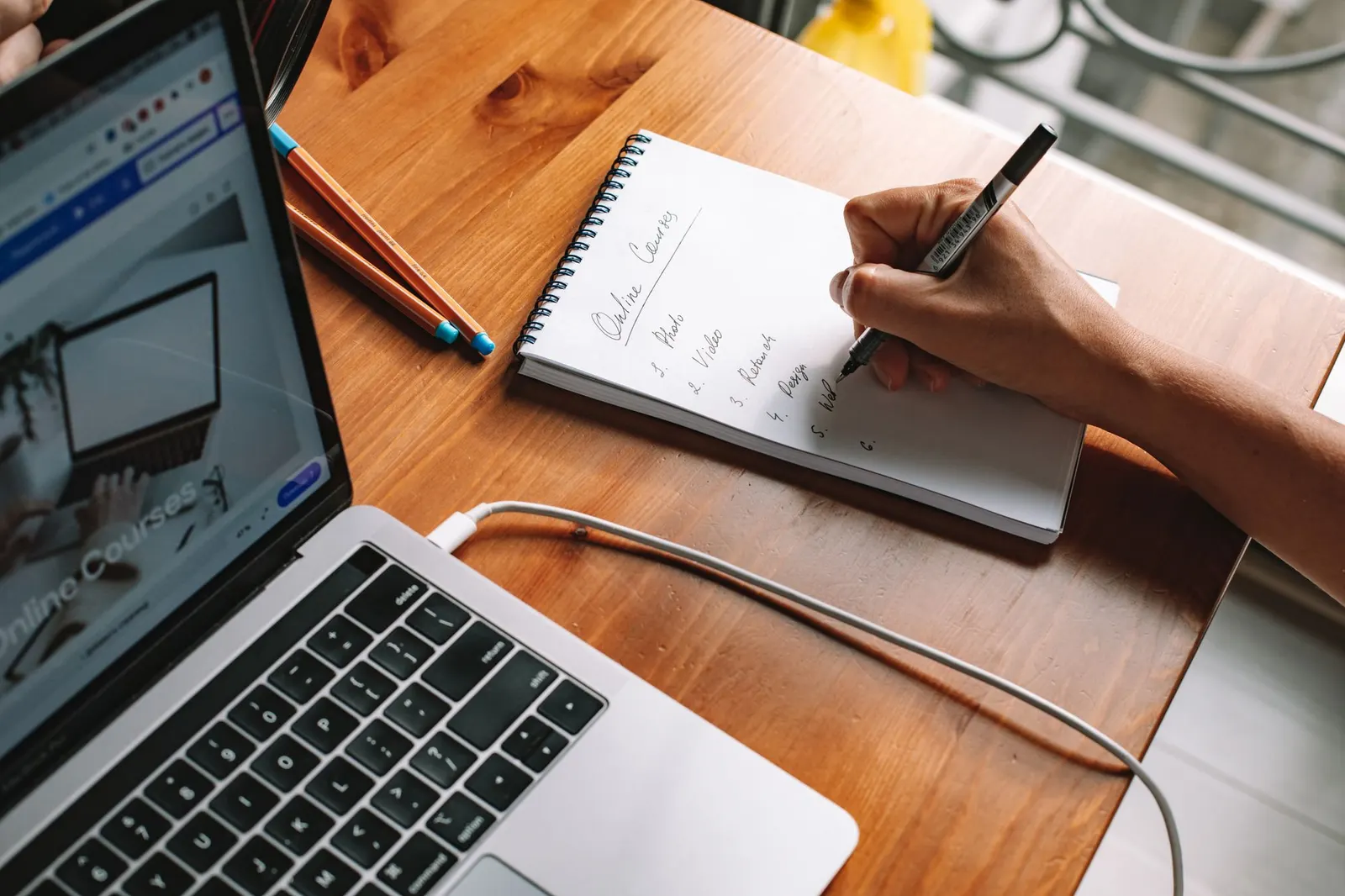Hand writing a numbered list in a notebook beside a laptop, studying Google AI Essentials