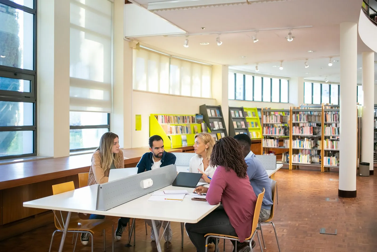 Mixed group of adult students talking around a table with laptops and notebooks in a bright, modern library