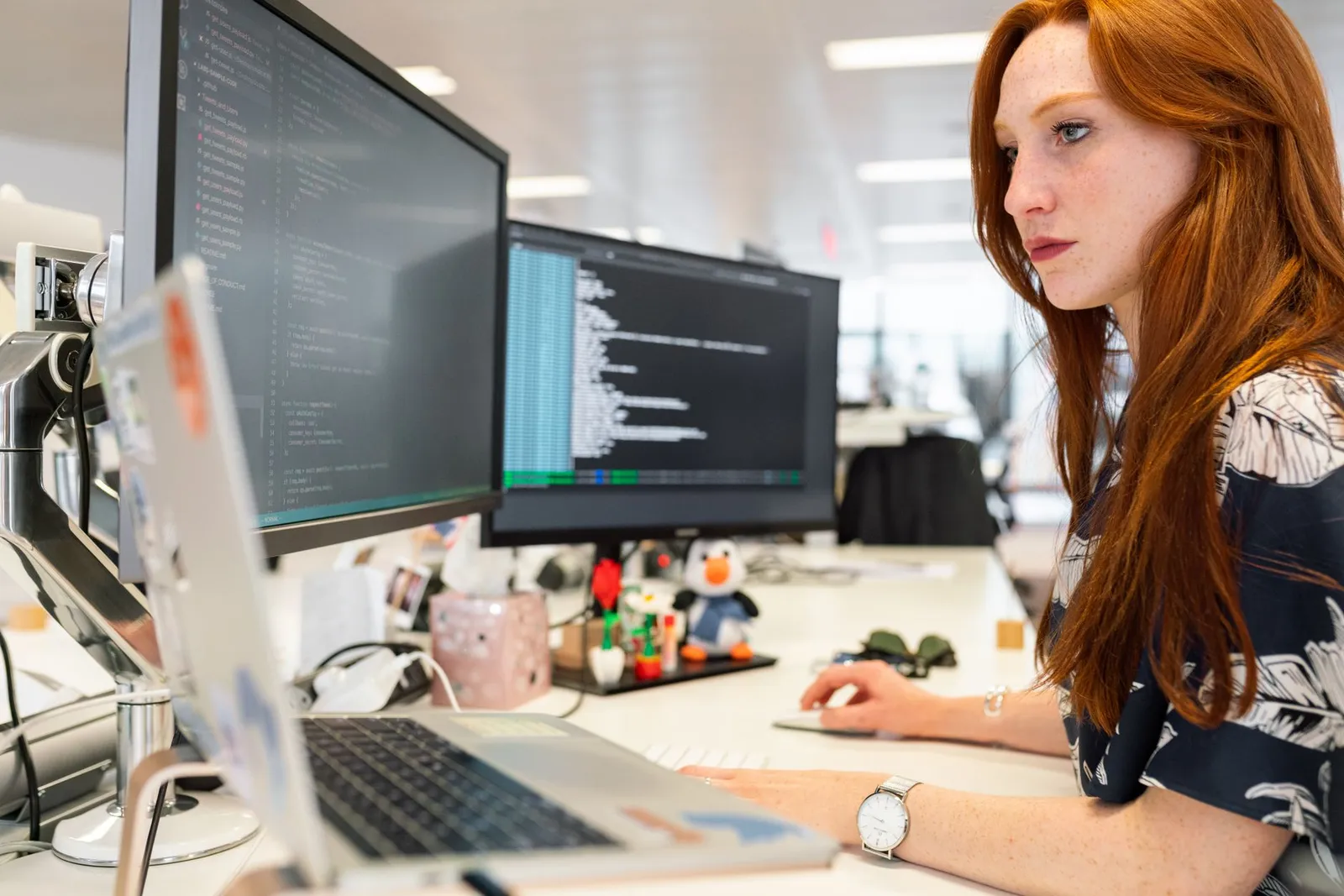 Machine learning engineer coding at a laptop and two monitors in an open office
