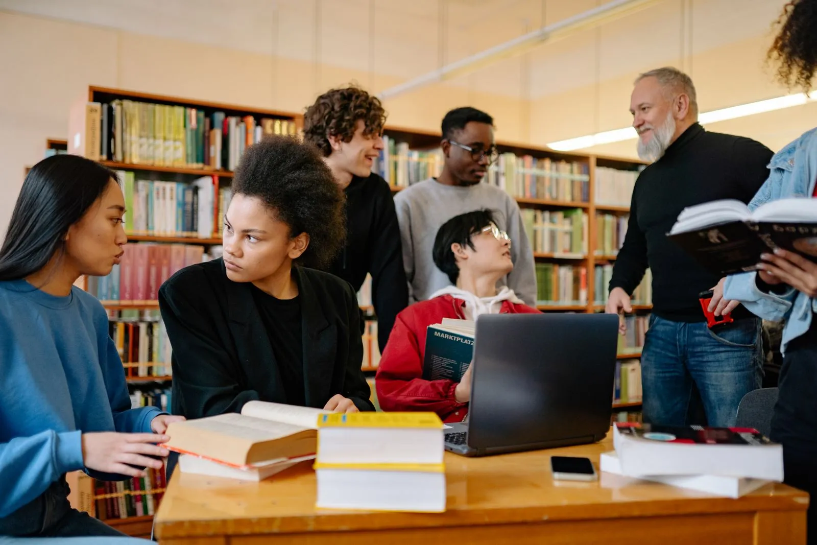 Students and an instructor discussing online AI degree rankings around a library table with books and a laptop