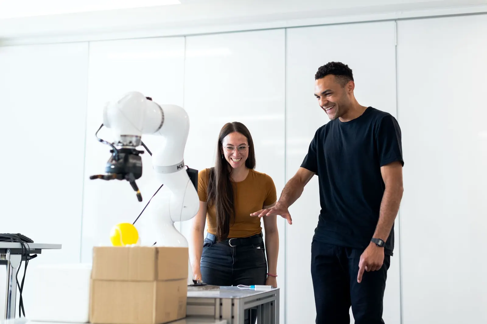 Robotics engineers smiling as a white robotic arm holds a gripper above a cardboard box