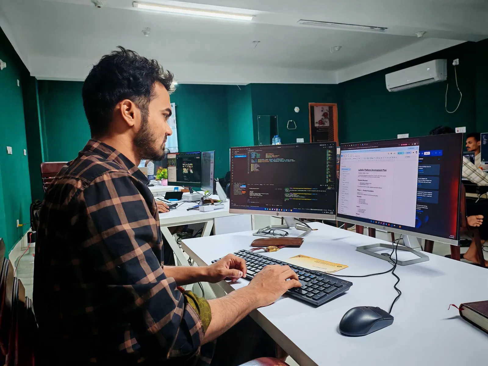 Software engineer typing at a white desk with two monitors showing code and a document